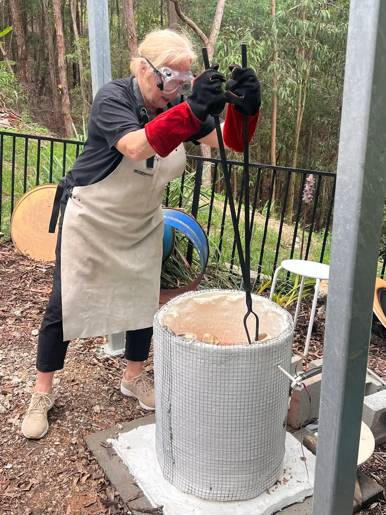 Deb Howells in gloves and goggles lifting the lid of a raku kiln with tongs